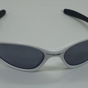 Puede incluir: Gafas de sol blancas con lentes grises oscuras y puntas de patillas negras. Las gafas tienen un dise&ntilde;o elegante y deportivo. El nombre de la marca es visible en la montura. Adecuadas para actividades al aire libre.