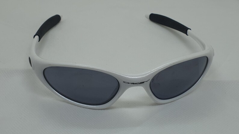 Puede incluir: Gafas de sol blancas con lentes grises oscuras y puntas de patillas negras. Las gafas tienen un dise&ntilde;o elegante y deportivo. El nombre de la marca es visible en la montura. Adecuadas para actividades al aire libre.