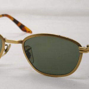 May include: Gold-framed sunglasses with green lenses. The frames have a rounded, oval shape and are paired with brown and black tortoise shell arms. The sunglasses are set against a white background.