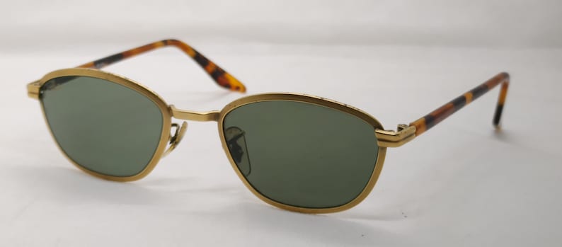 May include: Gold-framed sunglasses with green lenses. The frames have a rounded, oval shape and are paired with brown and black tortoise shell arms. The sunglasses are set against a white background.