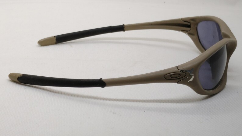 Puede incluir: Gafas de sol color beige con lentes oscuras. Las monturas son de color beige mate con detalles negros en las patillas. Las lentes son de color azul-gris oscuro. El logotipo de Oakley es visible en el lateral de la montura.