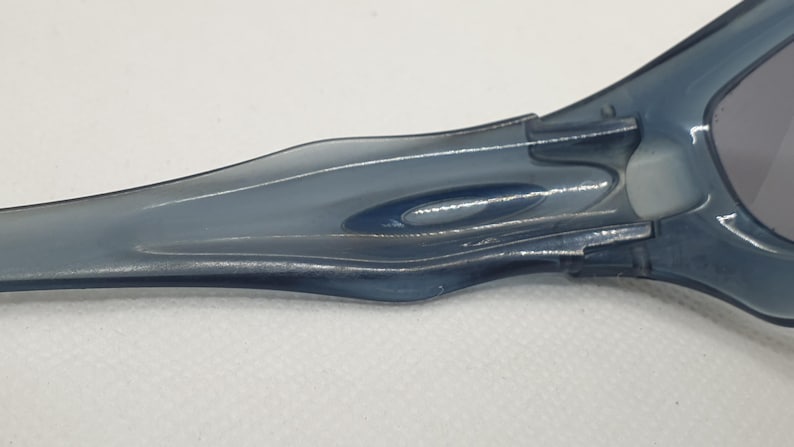 May include: Close-up of a pair of blue-tinted sunglasses. The image focuses on the arm and hinge of the glasses, showing the sleek, curved design and the connection point to the lens frame. The lenses are a dark gray color.