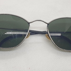 Puede incluir: Gafas de sol ovaladas con montura de metal plateado y lentes verde oscuro. Las patillas son negras y tienen un dise&ntilde;o curvado. Las lentes tienen algunos ara&ntilde;azos.