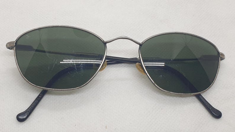 Puede incluir: Gafas de sol ovaladas con montura de metal plateado y lentes verde oscuro. Las patillas son negras y tienen un dise&ntilde;o curvado. Las lentes tienen algunos ara&ntilde;azos.
