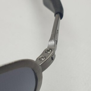 K&ouml;nnte beinhalten: Nahaufnahme einer Sonnenbrille. Der Rahmen ist mattgrau und die Gl&auml;ser sind dunkel. Das Scharnier und der B&uuml;gel der Sonnenbrille sind sichtbar, mit einem schwarzen Gummist&uuml;ck am Ende des B&uuml;gels.