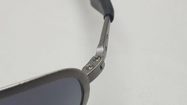 K&ouml;nnte beinhalten: Nahaufnahme einer Sonnenbrille. Der Rahmen ist mattgrau und die Gl&auml;ser sind dunkel. Das Scharnier und der B&uuml;gel der Sonnenbrille sind sichtbar, mit einem schwarzen Gummist&uuml;ck am Ende des B&uuml;gels.