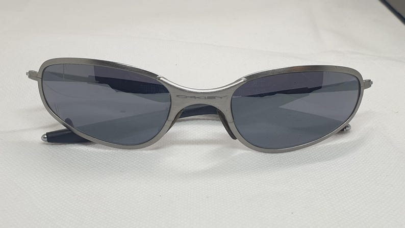 K&ouml;nnte beinhalten: Eine Sonnenbrille mit silbernem Rahmen und dunkelgrauen Gl&auml;sern. Die Sonnenbrille hat ein geschwungenes Design und der Markenname "Oakley" ist auf dem Steg sichtbar. Die B&uuml;gel sind schwarz.