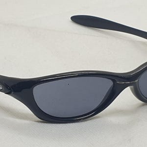 Puede incluir: Gafas de sol negras con un dise&ntilde;o elegante y curvado. Las lentes son de color gris oscuro y las monturas negras brillantes. Las patillas presentan un peque&ntilde;o logotipo plateado. Estas gafas ofrecen una est&eacute;tica moderna y deportiva.