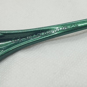 May include: Close-up of the arm of green sunglasses. The text "MADE IN U.S.A." is visible on the arm. The sunglasses have a metallic finish and a black arm tip.