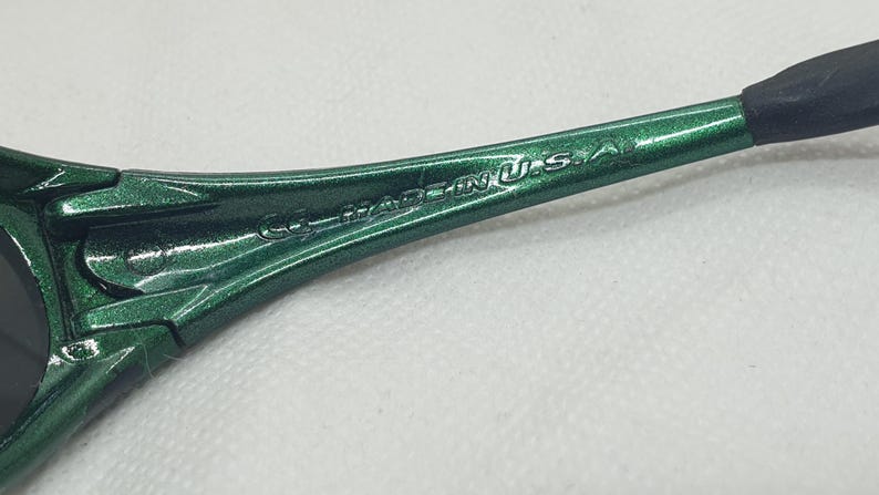 May include: Close-up of the arm of green sunglasses. The text "MADE IN U.S.A." is visible on the arm. The sunglasses have a metallic finish and a black arm tip.