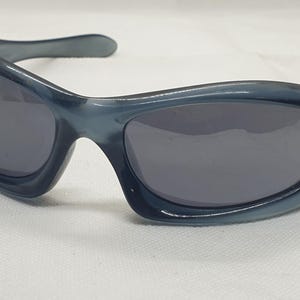 May include: Blue-tinted sunglasses with dark gray lenses. The sunglasses have a curved, wraparound design with a thick frame and arms. The lenses are oval-shaped, and the frame has a glossy finish. The sunglasses are on a white surface.