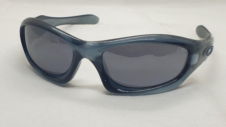 May include: Blue-tinted sunglasses with dark gray lenses. The sunglasses have a curved, wraparound design with a thick frame and arms. The lenses are oval-shaped, and the frame has a glossy finish. The sunglasses are on a white surface.