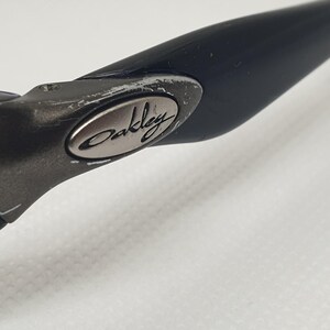 May include: Close-up of a black Oakley sunglass arm. The arm is made of a matte black material with a silver Oakley logo on the hinge. The hinge shows signs of wear. The background is a plain white surface.