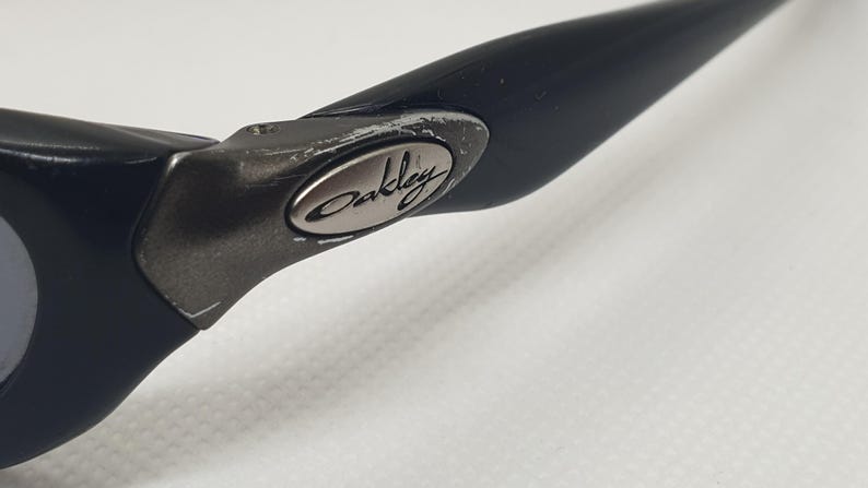 May include: Close-up of a black Oakley sunglass arm. The arm is made of a matte black material with a silver Oakley logo on the hinge. The hinge shows signs of wear. The background is a plain white surface.
