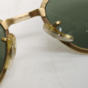 May include: Close-up of vintage sunglasses with gold-colored frames and green lenses. The frames show signs of wear, with a classic design. The lenses reflect a bright light source.