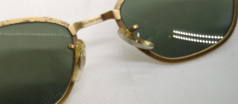 May include: Close-up of vintage sunglasses with gold-colored frames and green lenses. The frames show signs of wear, with a classic design. The lenses reflect a bright light source.