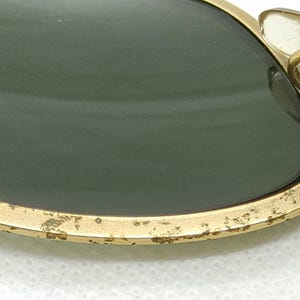May include: Close-up of vintage sunglasses with gold-colored frames and dark green lenses. The frame shows signs of wear, with some paint chipping. The brand name is visible on the lens and the temple arm.