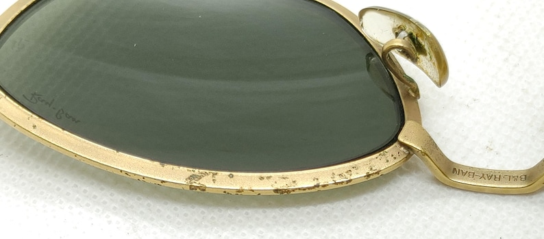 May include: Close-up of vintage sunglasses with gold-colored frames and dark green lenses. The frame shows signs of wear, with some paint chipping. The brand name is visible on the lens and the temple arm.
