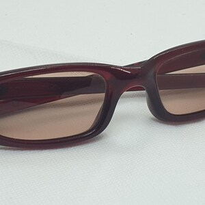 May include: Burgundy sunglasses with a rectangular frame and tinted lenses. The sunglasses have a distinctive design on the temples, set against a white background. A stylish accessory for eye protection.