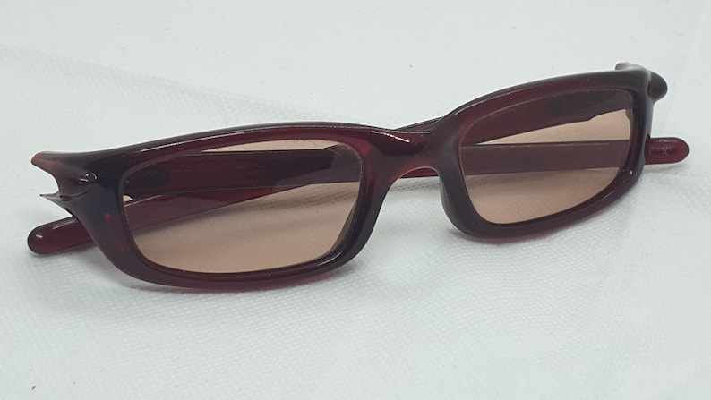 May include: Burgundy sunglasses with a rectangular frame and tinted lenses. The sunglasses have a distinctive design on the temples, set against a white background. A stylish accessory for eye protection.
