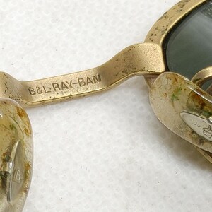 May include: Close-up of vintage gold-framed sunglasses with green lenses. The bridge of the glasses is marked with the text "B&L RAY-BAN". The nose pads are a light color. The sunglasses have a classic, timeless design.