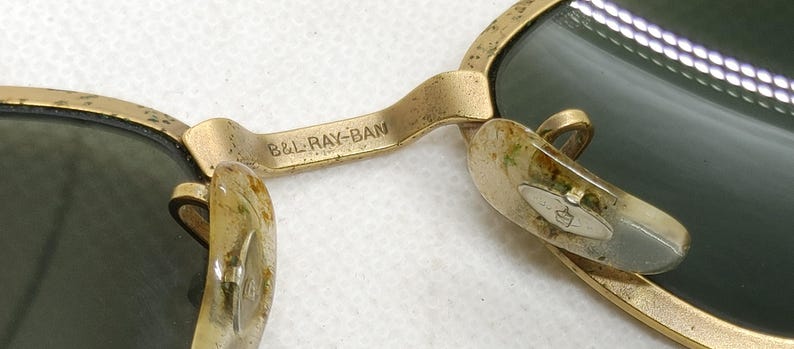May include: Close-up of vintage gold-framed sunglasses with green lenses. The bridge of the glasses is marked with the text "B&L RAY-BAN". The nose pads are a light color. The sunglasses have a classic, timeless design.