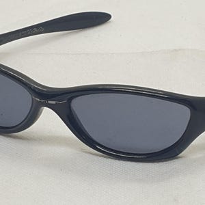 Puede incluir: Gafas de sol negras con montura curvada y lentes gris oscuro. Las gafas de sol tienen un peque&ntilde;o logotipo circular en las patillas. El dise&ntilde;o es elegante y moderno, adecuado para actividades al aire libre.