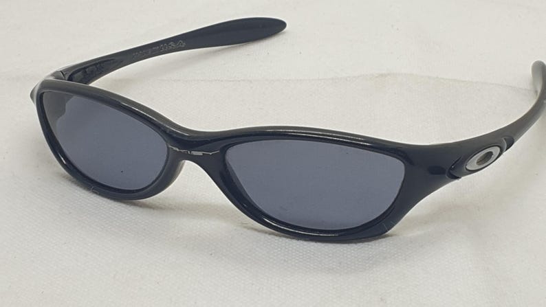 Puede incluir: Gafas de sol negras con montura curvada y lentes gris oscuro. Las gafas de sol tienen un peque&ntilde;o logotipo circular en las patillas. El dise&ntilde;o es elegante y moderno, adecuado para actividades al aire libre.