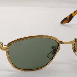 May include: Gold-rimmed sunglasses with oval green lenses. The temples feature a tortoiseshell pattern. The sunglasses are set against a white background. The frame has a classic, vintage design.