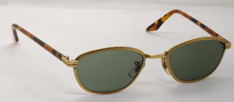 May include: Gold-rimmed sunglasses with oval green lenses. The temples feature a tortoiseshell pattern. The sunglasses are set against a white background. The frame has a classic, vintage design.