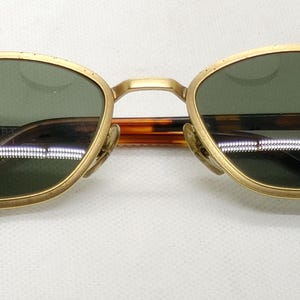 May include: Gold-framed sunglasses with green lenses and a tortoiseshell bridge and arms. The sunglasses have a classic, oval shape and are set against a white background. The brand name is visible on the lens.
