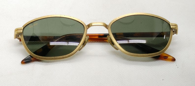 May include: Gold-framed sunglasses with green lenses and a tortoiseshell bridge and arms. The sunglasses have a classic, oval shape and are set against a white background. The brand name is visible on the lens.