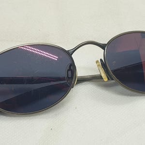 May include: Oval-shaped sunglasses with a dark metal frame and tinted lenses. The lenses have a gradient effect, transitioning from dark blue to a reddish hue. The arms of the glasses are thin and curve around the ears.