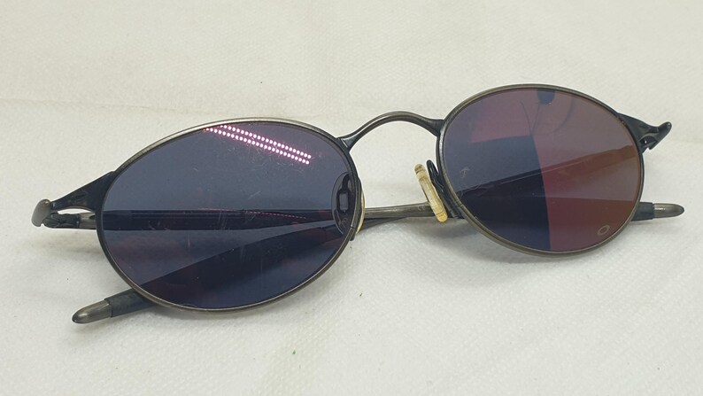 May include: Oval-shaped sunglasses with a dark metal frame and tinted lenses. The lenses have a gradient effect, transitioning from dark blue to a reddish hue. The arms of the glasses are thin and curve around the ears.