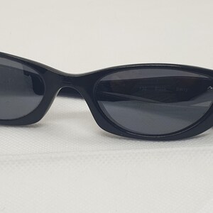 May include: Black sunglasses with a curved frame and dark lenses. The sunglasses have a sleek, modern design. The lenses are a dark grey colour, and the frame is a glossy black. The sunglasses are resting on a white surface.
