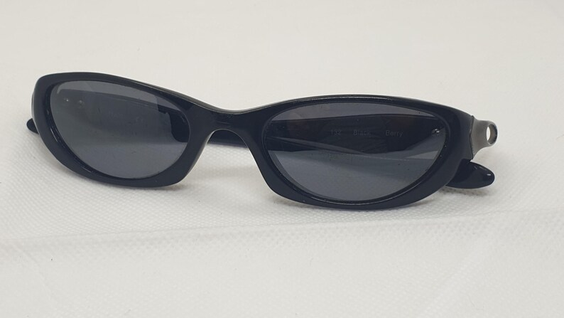 May include: Black sunglasses with a curved frame and dark lenses. The sunglasses have a sleek, modern design. The lenses are a dark grey colour, and the frame is a glossy black. The sunglasses are resting on a white surface.