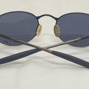 May include: Dark blue oval sunglasses with a thin, dark metal frame. The lenses are a matching dark blue. The temples are crossed, and the nose pads are gold-colored. The brand name is visible on the frame.