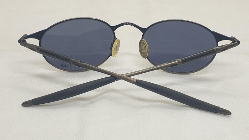 May include: Dark blue oval sunglasses with a thin, dark metal frame. The lenses are a matching dark blue. The temples are crossed, and the nose pads are gold-colored. The brand name is visible on the frame.