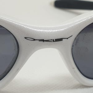 Puede incluir: Primer plano de gafas de sol Oakley blancas con lentes grises oscuras. El logotipo de Oakley est&aacute; impreso en negro en la montura. Las gafas de sol tienen un dise&ntilde;o deportivo y aerodin&aacute;mico, adecuado para actividades al aire libre.