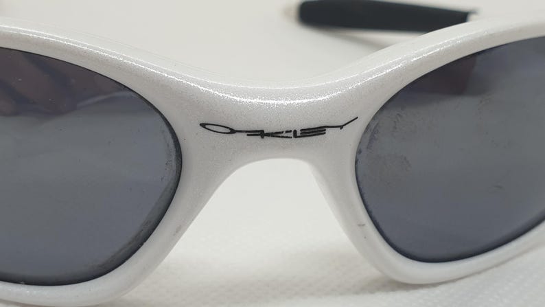 Puede incluir: Primer plano de gafas de sol Oakley blancas con lentes grises oscuras. El logotipo de Oakley est&aacute; impreso en negro en la montura. Las gafas de sol tienen un dise&ntilde;o deportivo y aerodin&aacute;mico, adecuado para actividades al aire libre.