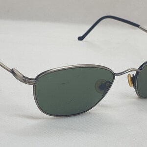 Puede incluir: Gafas de sol con montura plateada y lentes verdes ovaladas. Las monturas tienen un aspecto fino y met&aacute;lico con detalles negros en las patillas. Las gafas de sol est&aacute;n sobre un fondo blanco.