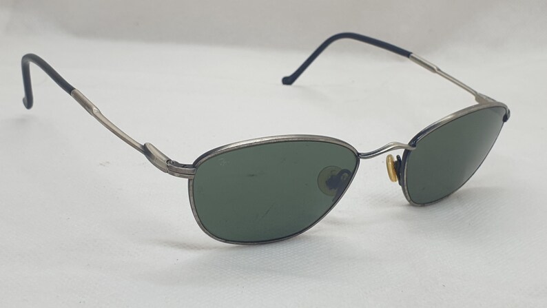 Puede incluir: Gafas de sol con montura plateada y lentes verdes ovaladas. Las monturas tienen un aspecto fino y met&aacute;lico con detalles negros en las patillas. Las gafas de sol est&aacute;n sobre un fondo blanco.