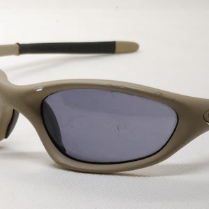 Puede incluir: Gafas de sol color beige con lentes oscuras. Las monturas tienen un dise&ntilde;o curvo con un acabado mate. Las patillas son negras con puntas beige. Las lentes son de color gris oscuro.
