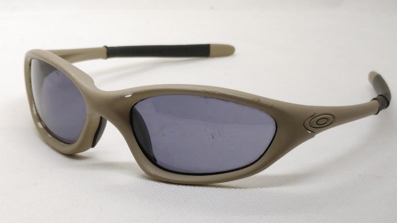 Puede incluir: Gafas de sol color beige con lentes oscuras. Las monturas tienen un dise&ntilde;o curvo con un acabado mate. Las patillas son negras con puntas beige. Las lentes son de color gris oscuro.