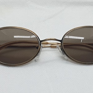 May include: Oval-shaped sunglasses with a bronze-colored frame and brown lenses. The temples and nose pads are a light gold color. The sunglasses are designed with a classic, vintage aesthetic.