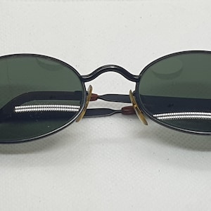 May include: Oval-shaped sunglasses with dark green lenses and a black metal frame. The temples are brown and the nose pads are gold. The sunglasses are set against a white background.