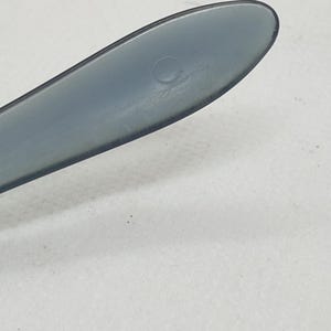 May include: Close-up of a gray plastic utensil, possibly a knife or spatula, with a rounded tip. The utensil has a smooth, matte finish and a small circular indentation near the tip. The background is a plain white surface.