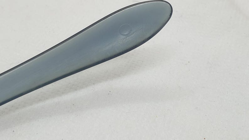 May include: Close-up of a gray plastic utensil, possibly a knife or spatula, with a rounded tip. The utensil has a smooth, matte finish and a small circular indentation near the tip. The background is a plain white surface.
