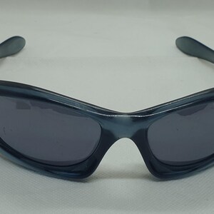 May include: Sport sunglasses with a wraparound design. The frames are a translucent blue-gray, and the lenses are a dark gray. The sunglasses have a sleek, modern aesthetic, suitable for outdoor activities and sports.