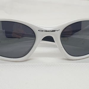 Puede incluir: Gafas de sol blancas de forma ovalada con lentes grises oscuras. Las gafas tienen una montura blanca con el nombre de la marca "Oakley" impreso en negro en el puente. Dise&ntilde;adas para la protecci&oacute;n ocular.